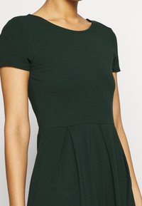 Abito corto a maniche corte di colore verde scuro con scollatura rotonda, caratterizzato da una parte inferiore plissettata e una texture di tessuto liscia.