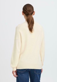 Cremfarbener Pullover mit gerippter Textur, entspannter Passform und tiefen Schultern. Kombiniert mit dunkelblauen Jeans, von hinten gezeigt.