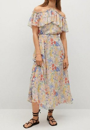 Vrouw draagt een off-shoulder bloemige midi-jurk met ruches bij de halslijn en zwarte platte veter-sandalen, staand tegen een effen achtergrond.