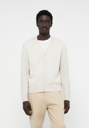 Lys beige cardigan laget av mykt stoff, med V-hals, lange ermer og seks knapper. Brukt over en hvit T-skjorte og beige bukser.