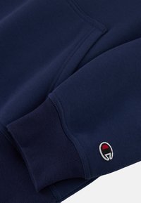 Dunkelblaues Sweatshirt mit weichem Obermaterial, ausgestattet mit einem gerippten Bündchen und einem kleinen gestickten Logo in Rot und Weiß am Ärmel.