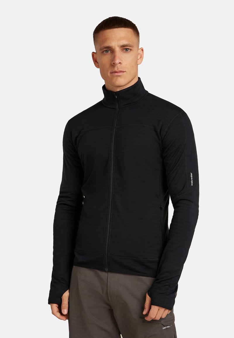Man in een zwarte ritsjack met hoge kraag en duimopeningen, gecombineerd met grijze broek, tegen een effen lichte achtergrond.