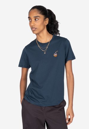 Frau mit dunklen Haaren im Pferdeschwanz, die ein dunkelblaues T-Shirt mit einem kleinen Ramen-Schüssel-Grafik und dunkle Hosen trägt, blickt nach links.