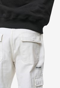 Hvite cargo bukser med to baklommer med klaff, stoffetikett med "PEGADOR"-logo, parret med en svart hettegenser med ribbestrikket kant.