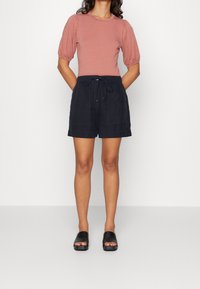 Light pink short-sleeve top with puffed shoulders, paired with dark navy linen shorts featuring an elastic waistband and drawstring. Black slide sandals.