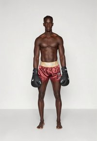 Boxeador com calções de cetim bordô com cintura elástica branca rotulada "VENUM", usando luvas de boxe pretas, em pé descalço.