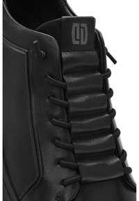 Zbliżenie na czarny skórzany trampek z żebrowanym językiem i czarnymi elastycznymi sznurowadłami przewleczonymi przez boczne przelotki.