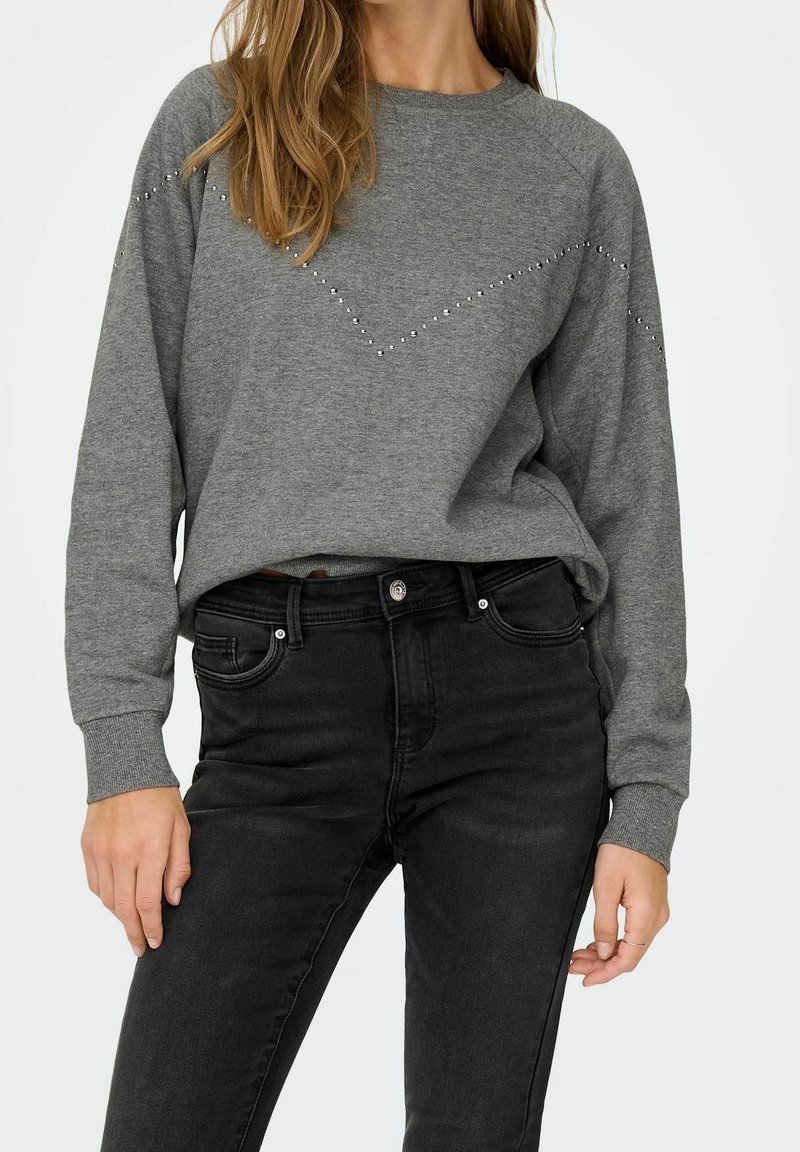 Femme portant un sweat-shirt gris à manches longues avec de petits clous en motif chevron et un jean noir taille haute.