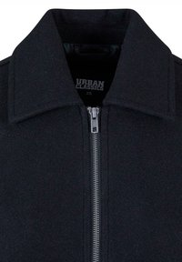 Veste noire zippée avec un large col, une fermeture éclair métallique, et une étiquette "Urban Classics XS" visible à l'intérieur du col.