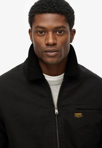 Veste en toile noire avec un col en velours côtelé, fermeture éclair et une petite étiquette orange. En dessous, il y a un pull léger gris.