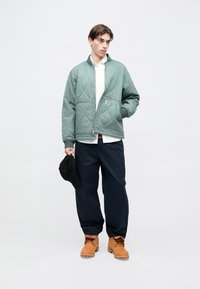 Jasnozielona pikowana kurtka z przednim zamkiem, biała koszula pod spodem, granatowe luźne spodnie oraz brązowe buty. Trzyma czarną czapkę.