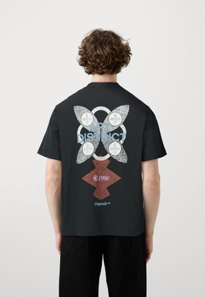 Personne aux cheveux bouclés portant un T-shirt noir avec un motif floral et les textes "ART DISTRICT," "©1990," et "Originals" au dos.
