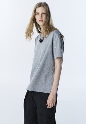 Giovane donna con lunghi capelli biondi che indossa una maglietta grigia oversize a pois, pantaloni neri a righe sottili e una collana nera spessa.