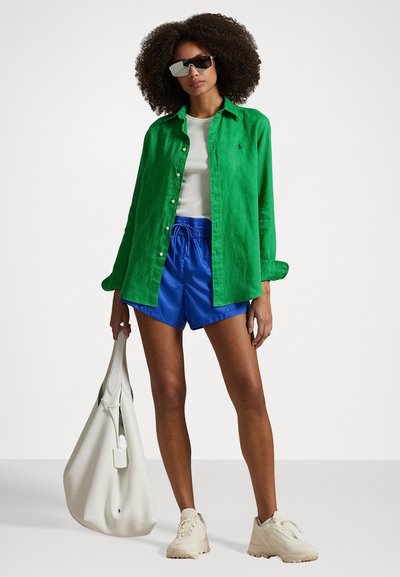 Cămașă verde din in cu nasturi, top alb, pantaloni scurți albaștri, geantă din piele albă și adidași bej. Accesoriile includ ochelari de soare argintii.
