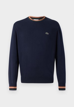 Sinih t-särk ümmarguse kaelusega, ribakäistega, millel on rohelised, oranžid ja valged triibud, ning väikese tikandiga logo rinnal.