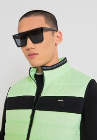 Occhiali da sole neri rettangolari e una giacca a vento verde menta con una striscia nera a coste e cerniere. Il colletto è a strisce bianche e nere.