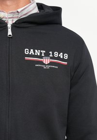 Crna zip-up dukserica s grafičkim printom na kojem piše "GANT 1949" i štitasti logo u crvenim i plavim akcentima. Mekani materijal s glatkom teksturom.