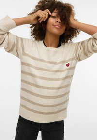 Pull beige tricoté avec des rayures marron clair, col rond et poignets côtelés. Comprend un petit cœur rouge brodé sur la poitrine.