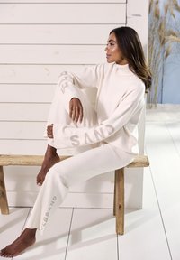 Witte, gebreide lounge-outfit bestaande uit een trui met hoge kraag en wijde broek, met de tekst "SAND" in een gedempte kleur op de mouwen en benen.