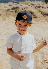 Fekete snapback sapka bézs emblémával, fehér, rövid ujjú gombos ing, homokos talajon állva, egy kis ágat tartva.