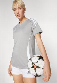 Grå atletisk skjorte med hvite detaljer, kombinert med hvite shorts. Spilleren holder en mønstrede svart og hvit fotballs med oransje detaljer.