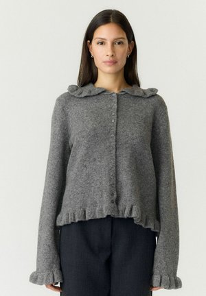Donna che indossa un maglione grigio abbottonato con collo, orlo e polsini arricciati, abbinato a pantaloni scuri, in piedi davanti a uno sfondo uniforme.