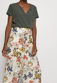 Haut vert foncé à manches courtes avec un décolleté en V et une poche poitrine, associé à un pantalon floral à jambe large en crème, avec des motifs rouges, jaunes et bleus.