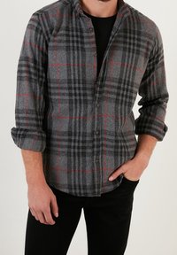Camicia di flanella grigia con motivo a quadri neri e rossi, con bottoni sul davanti, maniche arrotolate e orlo leggermente curvato. Indossata sopra una maglietta nera.