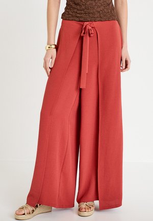 Femme portant un pantalon large ceinturé de couleur rouille, avec une taille nouée, et des sandales compensées dorées, debout devant un fond blanc uni.