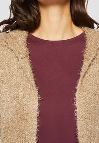 Beige neulottu neuletakki pörröisellä pinnalla, avoin edestä. Päällä tummanpunainen, pyöreäkauluksinen paita. Hiukset osittain näkyvissä.