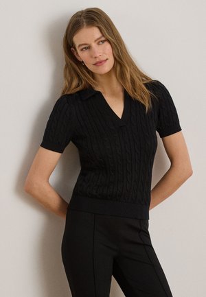Femme aux longs cheveux châtains clairs portant un pull noir à manches courtes en maille torsadée avec col, debout devant un mur clair uni.