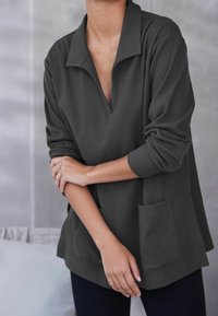 Pullover gris foncé à côtes avec un col en V et des manches longues, doté d'une poche avant et d'un design ample. Matériau doux et texturé.