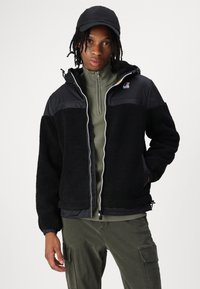 Schwarze Fleecejacke mit einem glänzenden schwarzen Oberteil, durchgehendem Reißverschluss, Kordelzug am Saum, getragen über einem olivgrünen Reißverschluss-Pullover. Schwarze Mütze auf dem Modell.