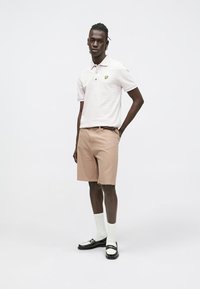 Witte polo met een geel logo, gecombineerd met beige korte broek met krijtstrepen. Heeft zwarte schoenen en witte enkelsokken. Eenvoudig, strak ontwerp.