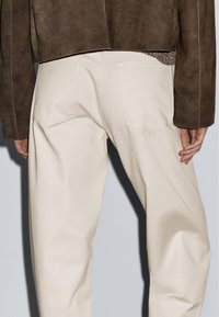 Hnědá semišová bunda s kratším střihem, spárovaná se světlými béžovými kalhotami s širokými nohavicemi a zadními kapsami, vyrobenými z hladkého bavlněného materiálu.