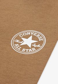Brūns audekls ar baltu "Converse All Star" logotipu, kurā ir zvaigzne apļa malā. Teksts un detaļas ir skaidri definētas un izteiksmīgas.