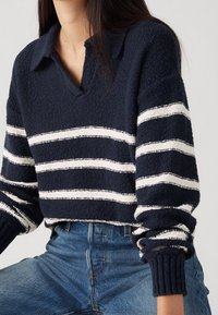 Tmavě modrý pletený svetr s výstřihem do V a dlouhými rukávy, zdobený bílými vodorovnými pruhy. Strukturální tkanina s žebrovanými lemy na rukávech.