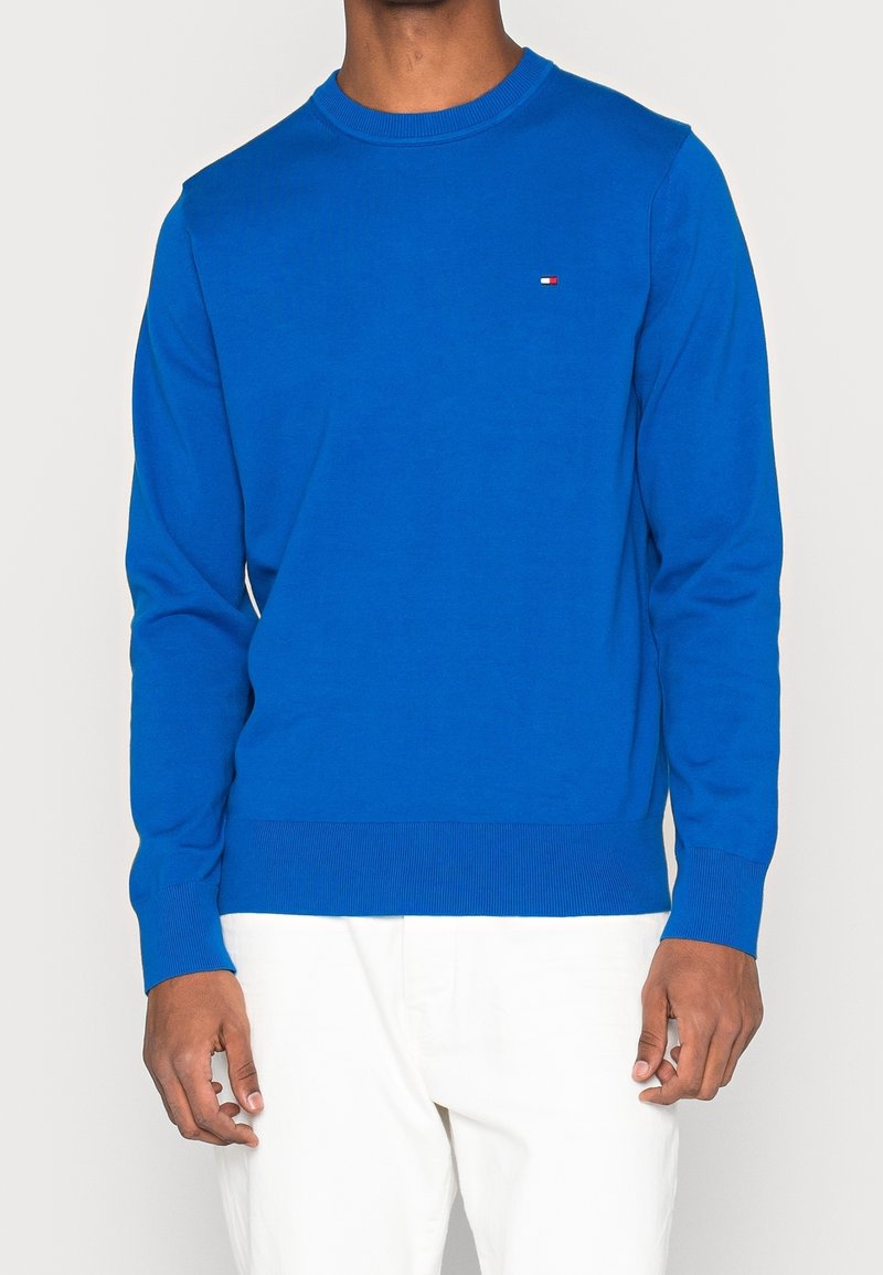 Homme portant un pull bleu vif à manches longues avec un petit logo rectangulaire sur la poitrine, assorti à un pantalon blanc.