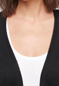 Black knitted cardigan over a white tank top, featuring a v-neckline and textured fabric. The focus is on the layering of the garments.