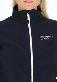 Navyfärgad fleecejacka med hög krage och vit dragkedja. Har "SWEDEMOUNT"-logotypen broderad på vänster sida. Slät textur.