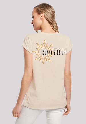 Beige Kurzarm-T-Shirt mit einem Sonnen-Design und dem Text „HALTE DEINE SONNENSEITE NACH OBEN“ auf dem Rücken. Weicher Stoff, entspannte Passform und glatte Textur.