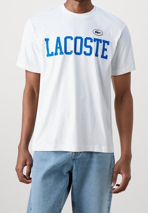 Valkoinen puuvillainen t-paita, jossa on rohkea sininen "LACOSTE"-logo, pyöreä pääntie, lyhyet hihat ja pieni Lacoste-krokotiiliembleemi rinnassa.