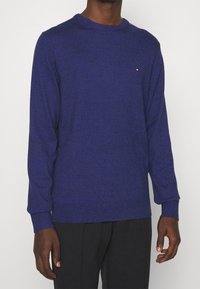 Maglione viola a girocollo con maniche lunghe, realizzato in morbido tessuto a maglia. Presenta un piccolo logo sul petto e polsini e orlo a coste.