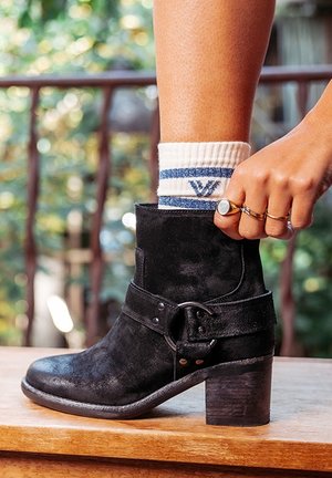 Bottine en daim noir avec un talon épais, dotée d'une bride à boucle et d'une finition texturée. Portée avec une chaussette blanche et des bagues argentées.