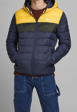 Mann trägt eine Kapuzen-Steppjacke in Gelb, Dunkelgrün und Navyblau mit den Händen in den Taschen, kombiniert mit blauen Jeans.