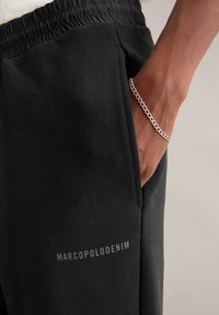 Schwarze Sweatpants mit elastischem Bund, ausgestattet mit einer Seitentasche und dem in Grau auf dem Oberschenkel gedruckten "MARCOPOLODENIM"-Logo. Sichtbare silberne Kette.