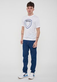T-shirt bianca a maniche corte con logo a scudo blu, abbinata a jeans blu scuro e scarpe da ginnastica bianche con accenti blu. Design semplice e casual.