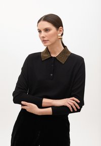 Schwarzes Langarm-Polohemd mit einem genietenen goldenen Kragen, ausgestattet mit einer Knopfleiste und gerippten Bündchen. Kombiniert mit schwarzen strukturierten Hosen.