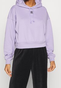 Lavendelfarbene cropped Hoodie mit Kordelzugkapuze, gerippten Bündchen und kleinem schwarzem Logo auf der Brust, kombiniert mit schwarzen Cordhosen.