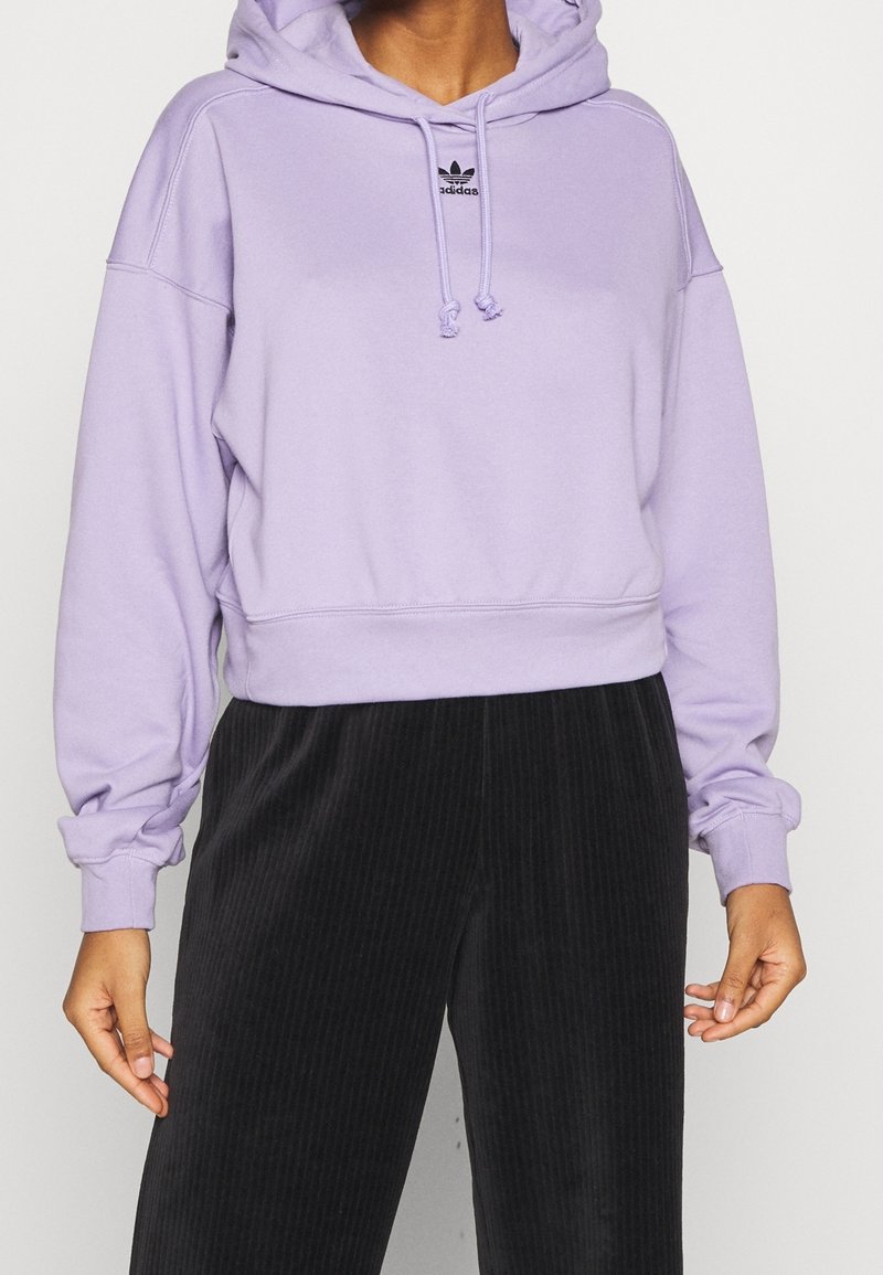 Lavendelfarbene cropped Hoodie mit Kordelzugkapuze, gerippten Bündchen und kleinem schwarzem Logo auf der Brust, kombiniert mit schwarzen Cordhosen.